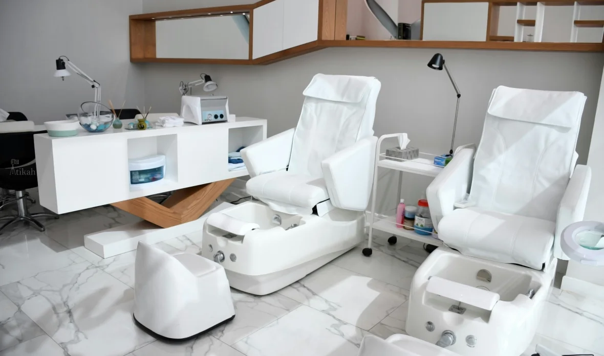 Ein heller Kosmetikbereich mit zwei weißen Pediküre-Sesseln, Fußbädern und einem weißen Behandlungstisch mit Pflegeutensilien auf glänzendem Marmorboden.